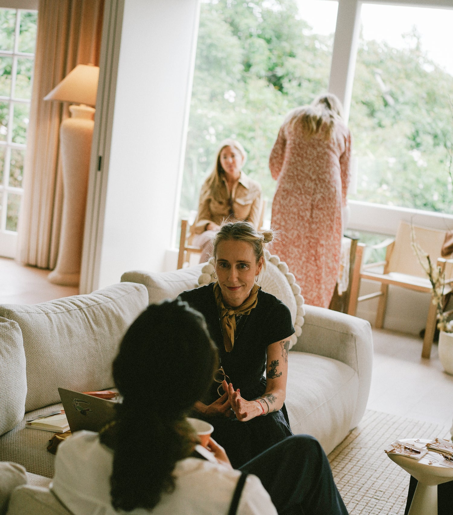 Woman sitting on a couch in a living room with another person, surrounded by large windows and indoor plants.